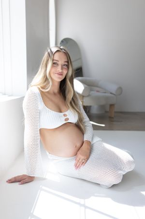 Expectant mother in white crochet outfit sits in bright minimalist room.