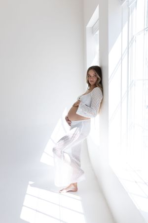 Person in white outfit stands by bright window in minimalist setting.