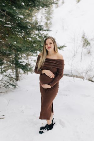 Person in brown dress poses in snowy winter scene with evergreen trees.