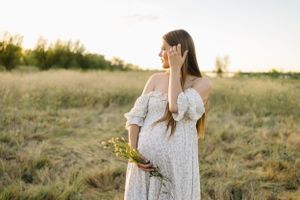 Maternity portrait of woman in white dress holding wildflowers in golden field.
