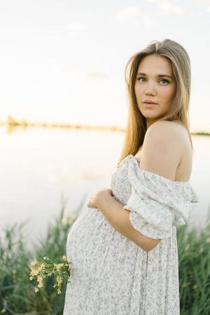 Expectant mother in white lace dress poses at sunset by water.