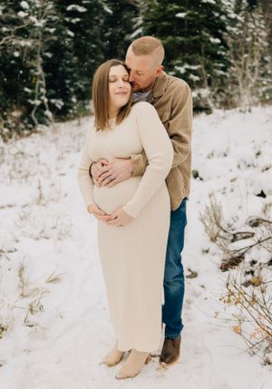 Intimate winter maternity photo with couple embracing in snowy forest.