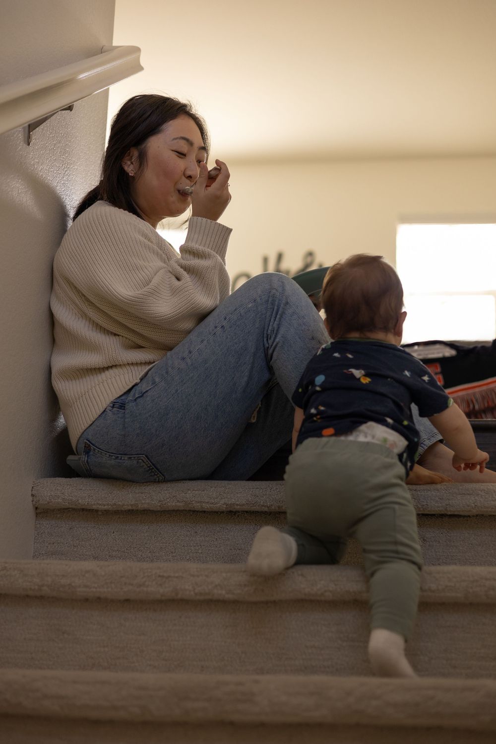 Mom eating at the top of the stairs, smiling at toddler boy climbing up the stairs curiously in a cozy in-home session in Murrieta.