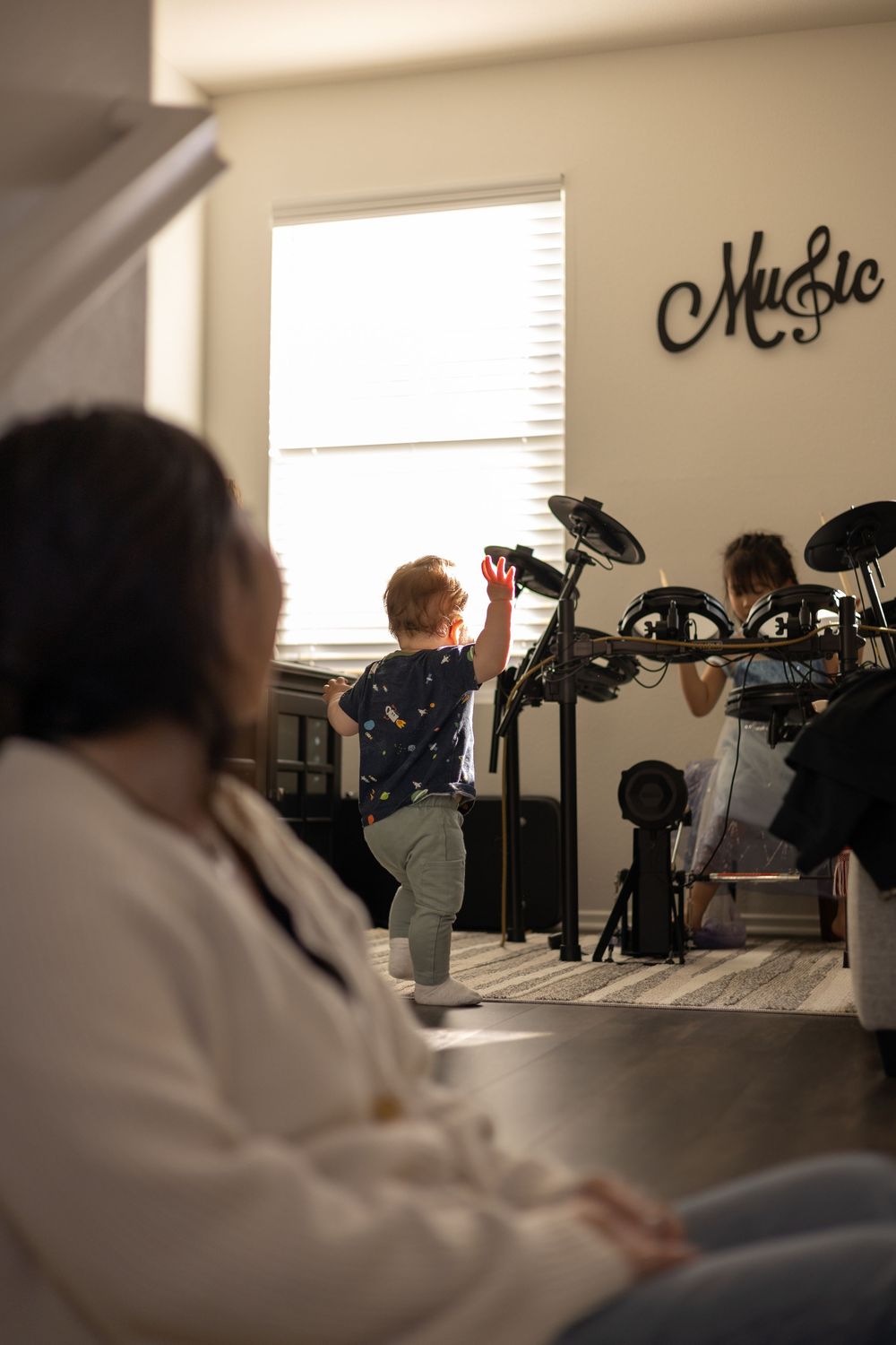 Mother watching toddler son dance next to drum set played by young girl during cozy, in-home motherhood session in Murrieta, CA.