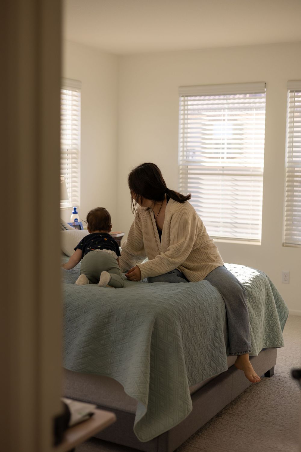 Distant shot of mom and toddler boy sitting on the bed in a warmly lit room in Murrieta.