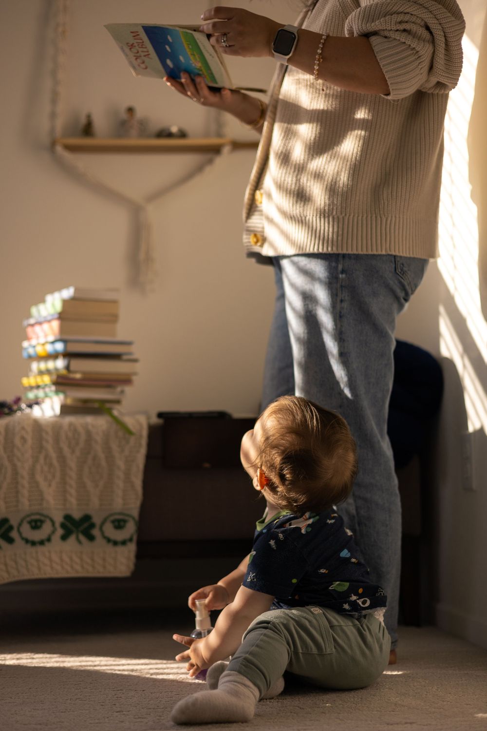 In-home session of mom looking at album and toddler boy sitting at her feet looking up curiously at her, with warm dappled light, in Murriet