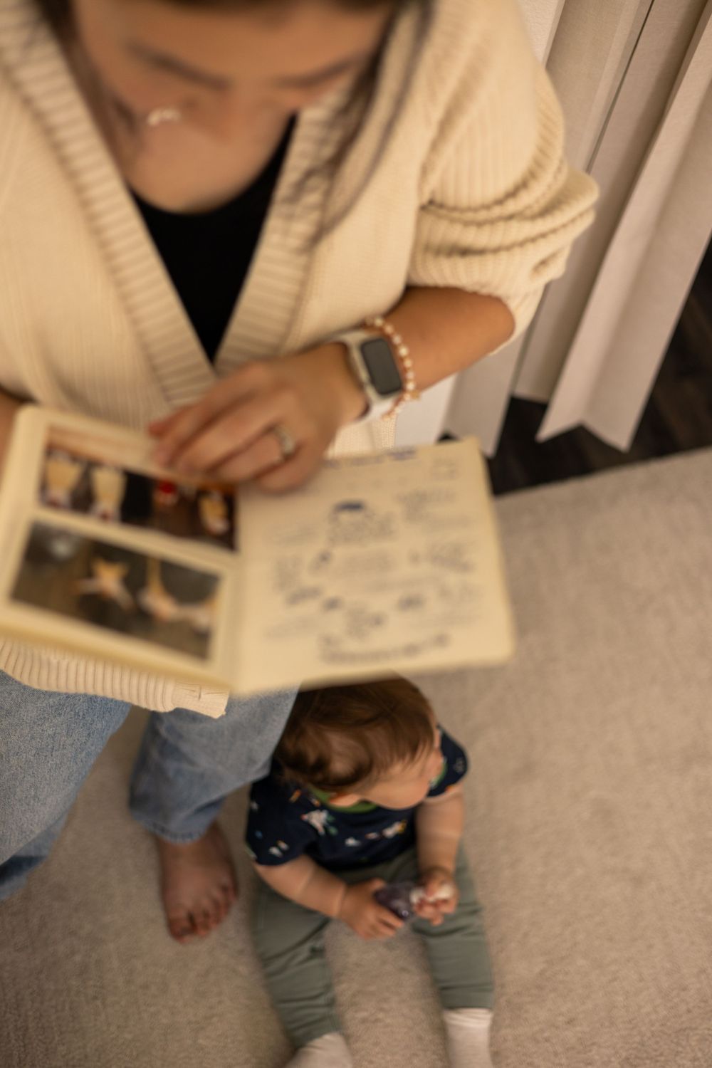 Nostalgic overhead shot of mom looking through an old album, standing up, as toddler boy sits at her feet looking to the side in Murrieta.