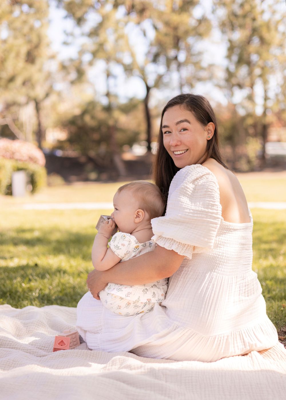 Both mom and baby girl are sitting on the picnic blanket, but mom is looking back at camera and smiling in San Diego.