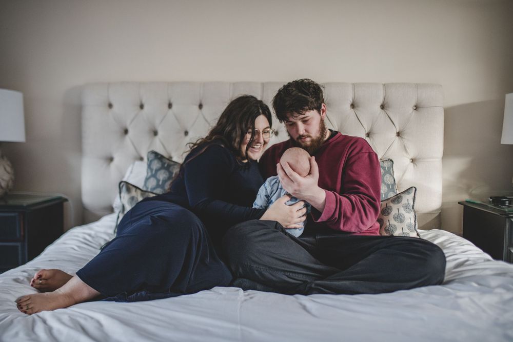 Couple sitting on gray upholstered bed while snuggling with newborn baby in cozy bedroom.