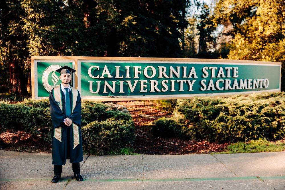Graduate in cap and gown standing in front of California State University Sacramento entrance sign in fall setting.