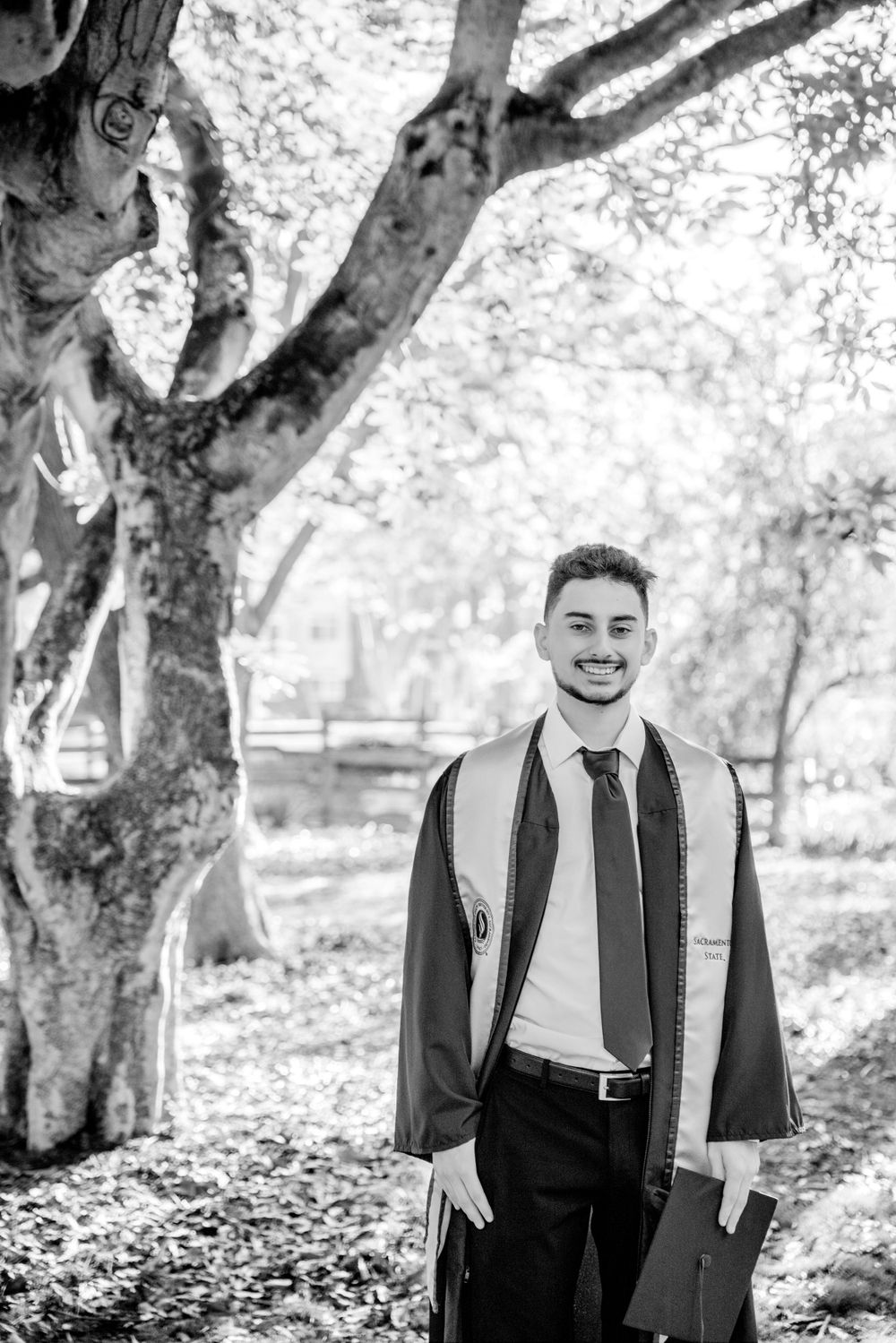 Black and white graduation portrait taken outdoors under shady trees showing graduate in academic regalia holding diploma.