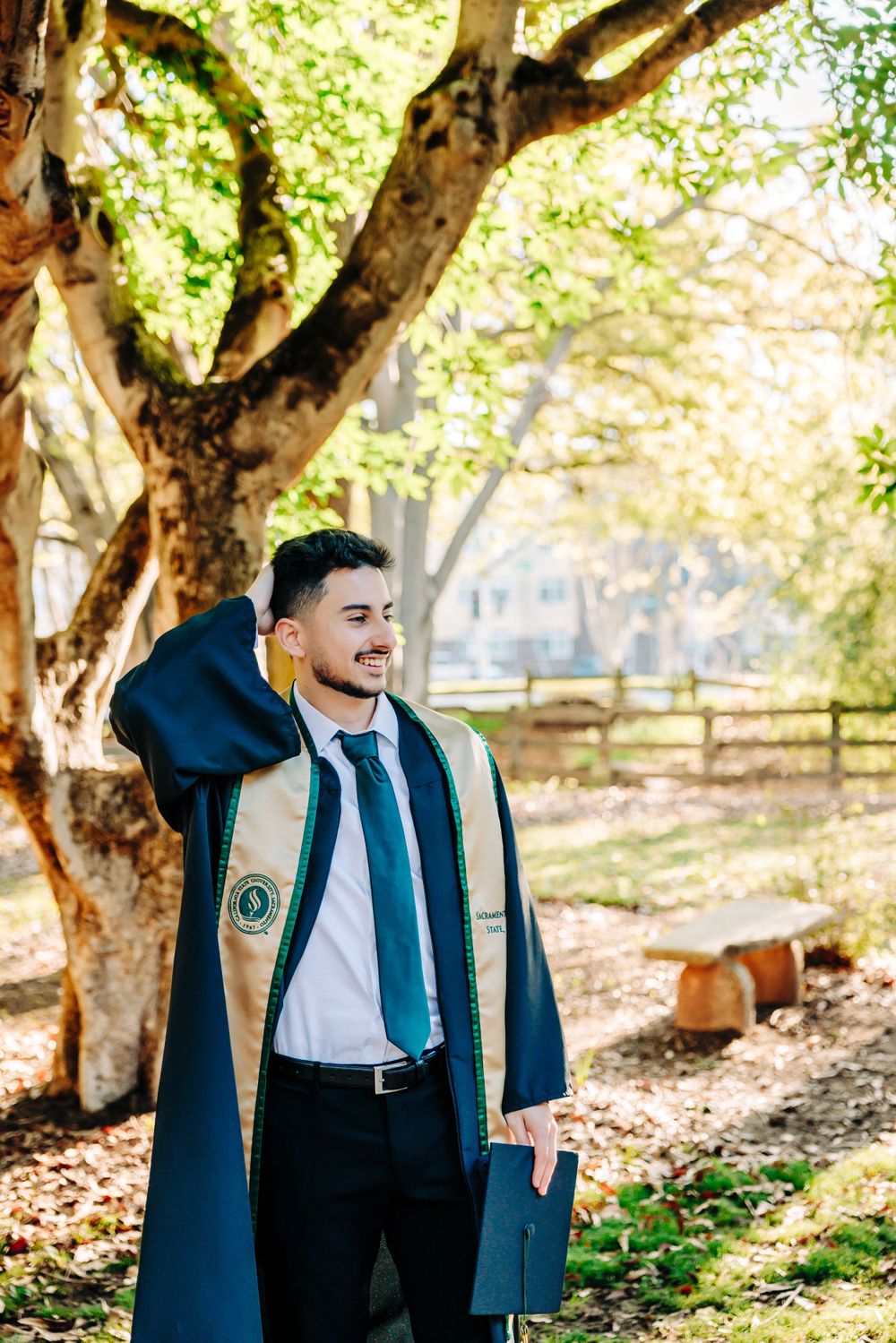 Graduate posing outdoors in blue academic regalia against autumn trees and natural lighting.