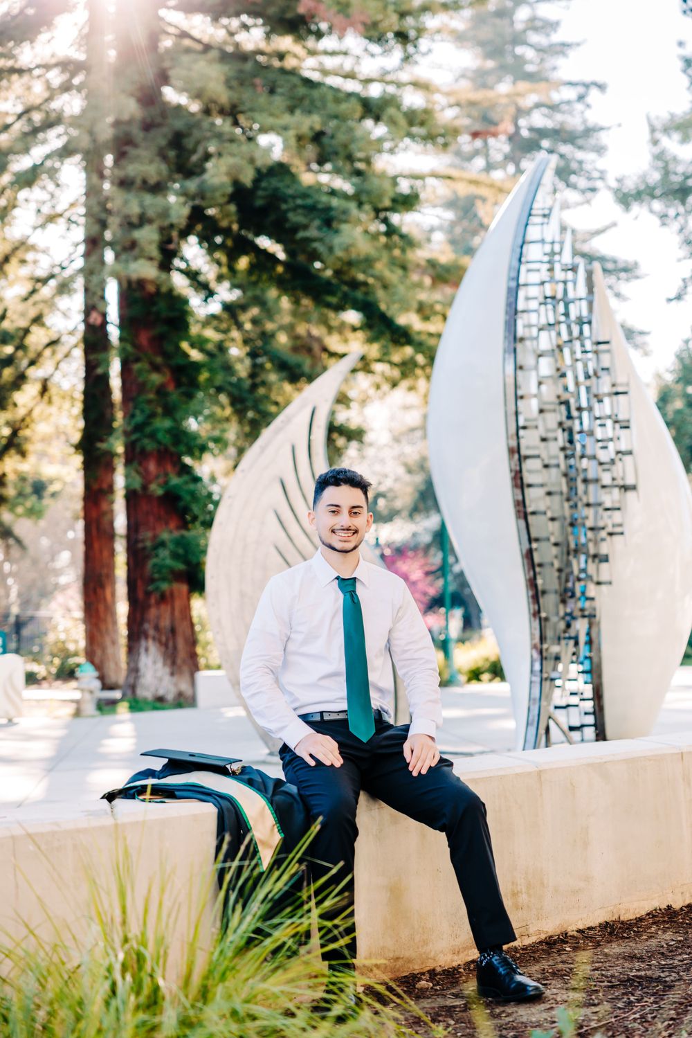 Professional portrait in front of modern metal sculpture with sunlit trees in background on university campus.