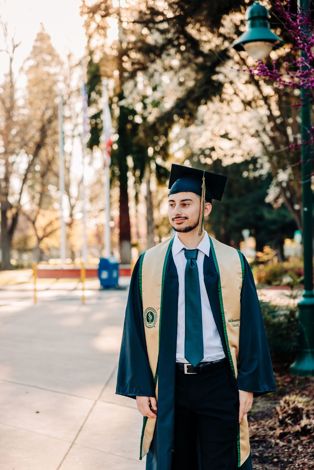 Graduate in traditional cap and gown with gold stoles stands on campus pathway during golden hour.