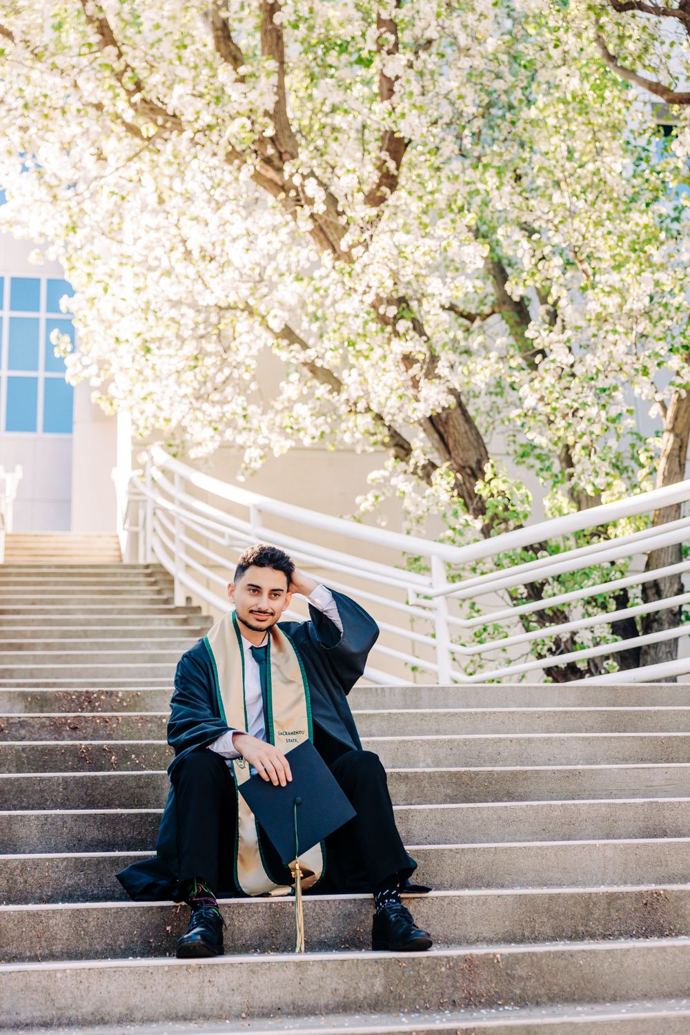 Graduate in cap and gown sitting on concrete stairs with blooming cherry blossoms overhead on sunny spring day.