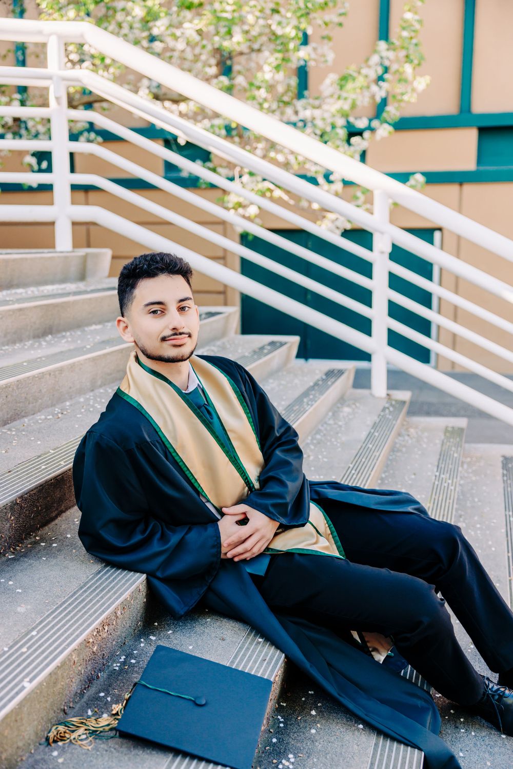 A graduate in traditional navy blue and gold regalia sits on concrete steps with their graduation cap nearby.