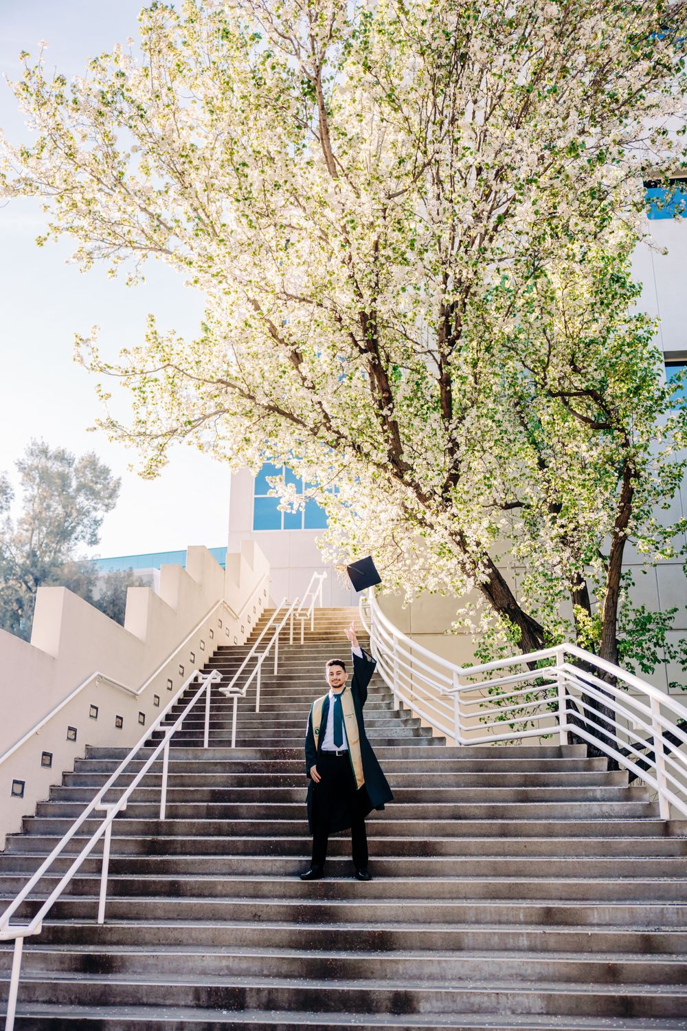 Graduate throws graduation cap in celebration on outdoor staircase surrounded by spring blossoms.