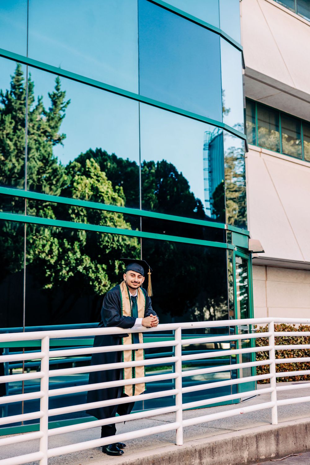 Graduate in cap and gown poses outside modern glass office building with reflective windows and white railings.