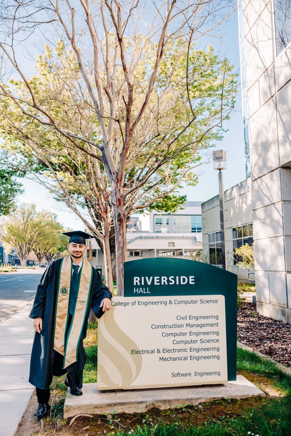 Graduate in cap and gown poses next to Riverside Hall engineering department sign on sunny college campus.