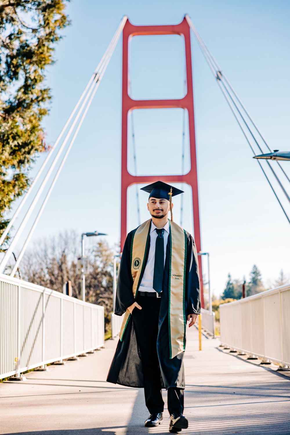 A graduate in cap and gown poses on a pedestrian bridge with the iconic red Golden Gate Bridge towers in the background.