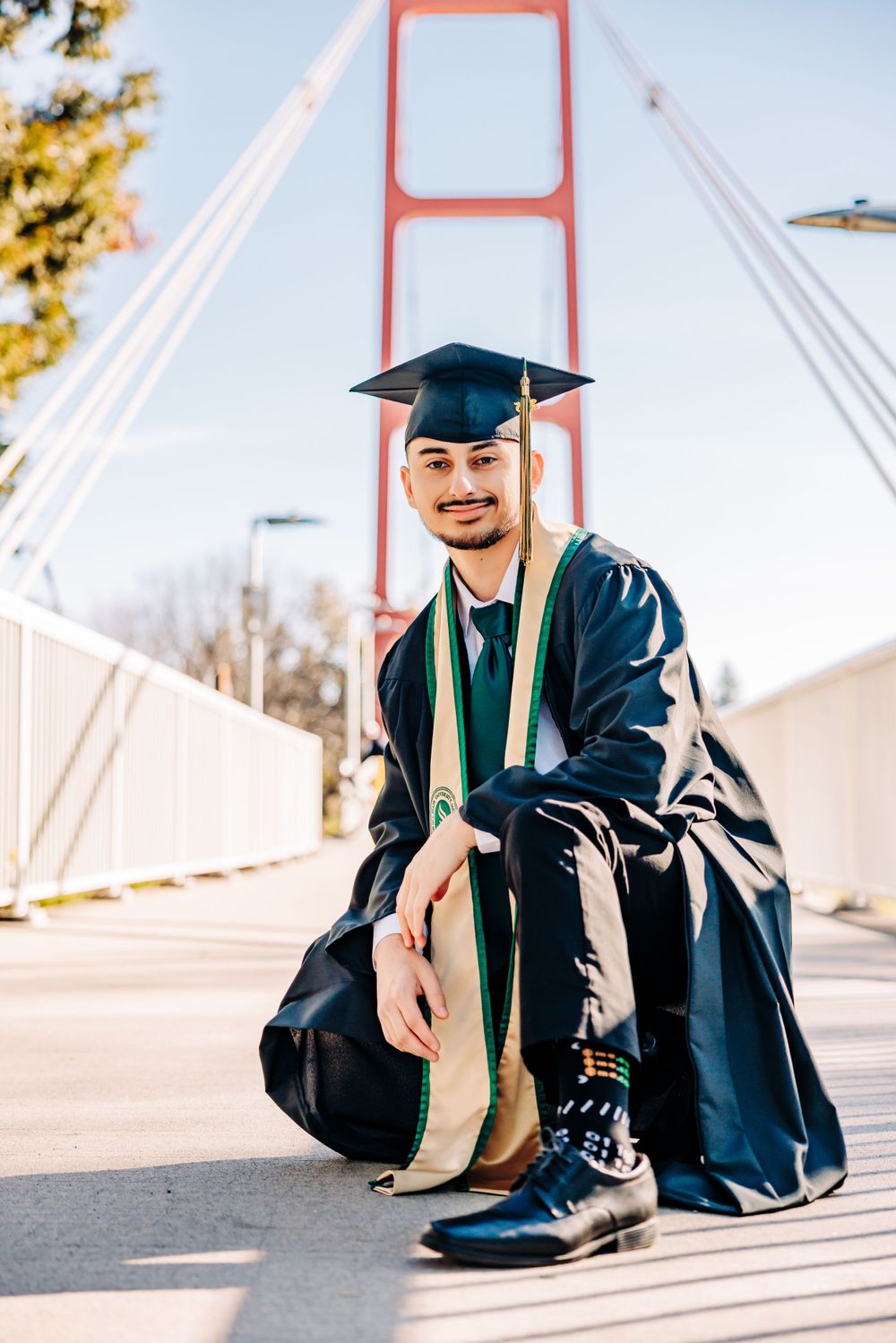 Graduate in cap and gown posing on a bridge with distinctive red architectural elements.