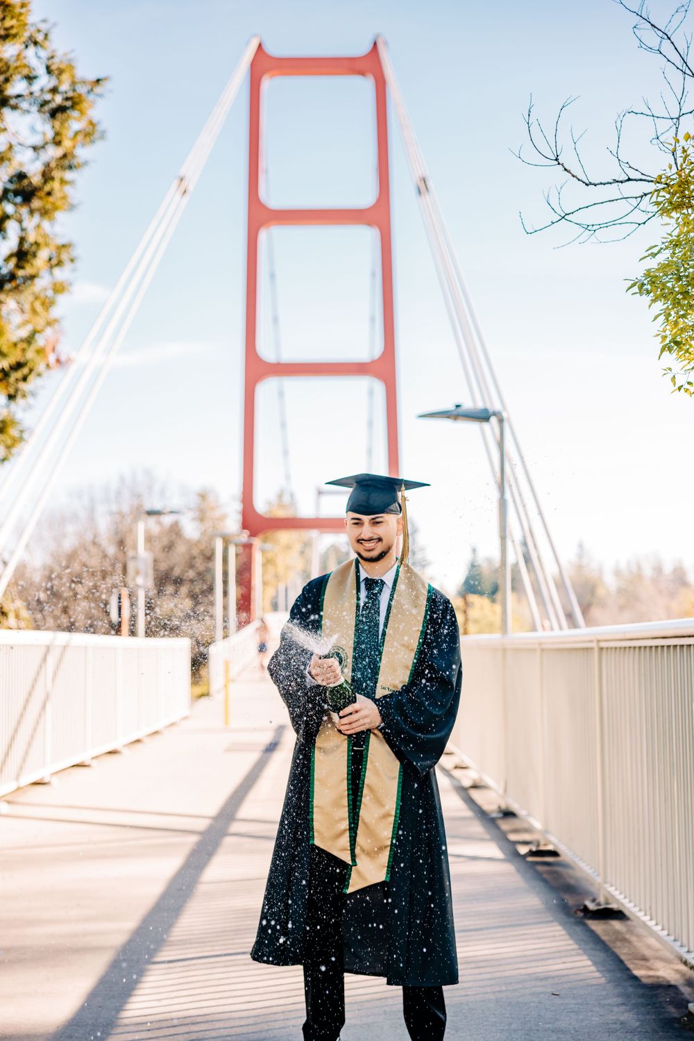 A graduate in traditional cap and gown poses on a bridge with a red suspension tower in the background.