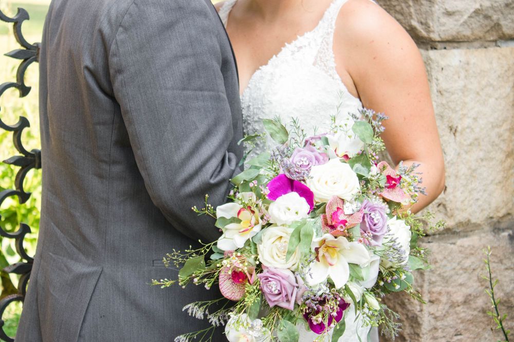 A colorful wedding bouquet featuring pink and white flowers with greenery held between a gray suit and white dress.