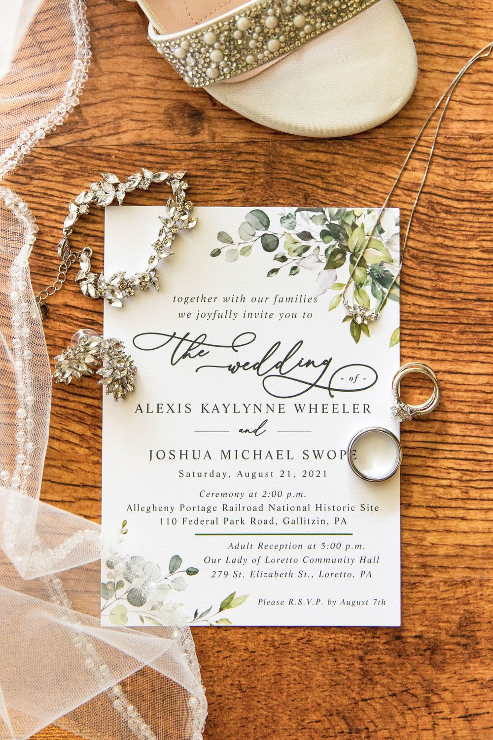 Wedding invitation with floral design displayed with rings, jewelry and elegant bridal shoes on wooden surface.