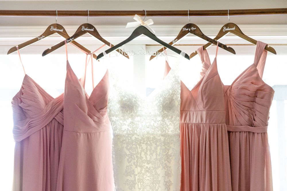 Wedding gowns in blush pink and ivory hang on wooden hangers against a white wall, creating an elegant bridal display.