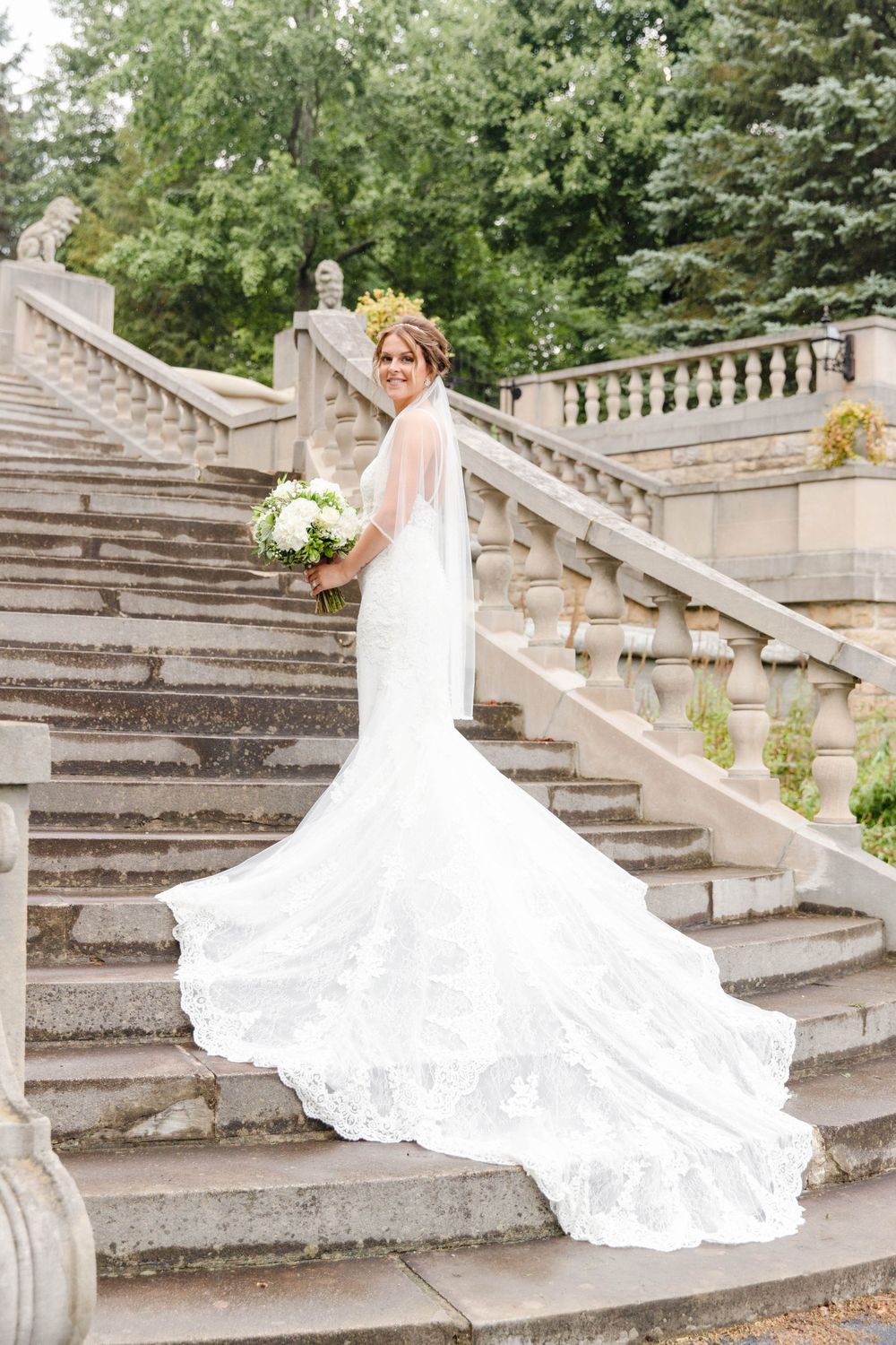 Beautiful wedding dress with long lace train displayed on elegant stone staircase in outdoor garden setting.