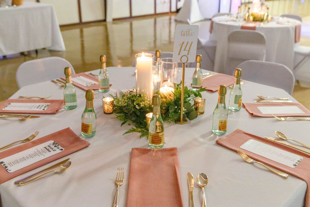 Elegant wedding table setting with blush napkins, candles, and greenery centerpiece on white tablecloths.