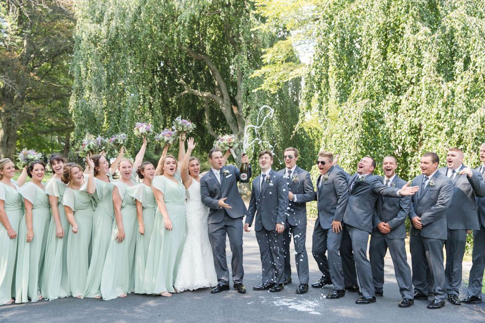 Wedding party in mint green dresses and navy suits pose together under weeping willow trees in a garden setting.