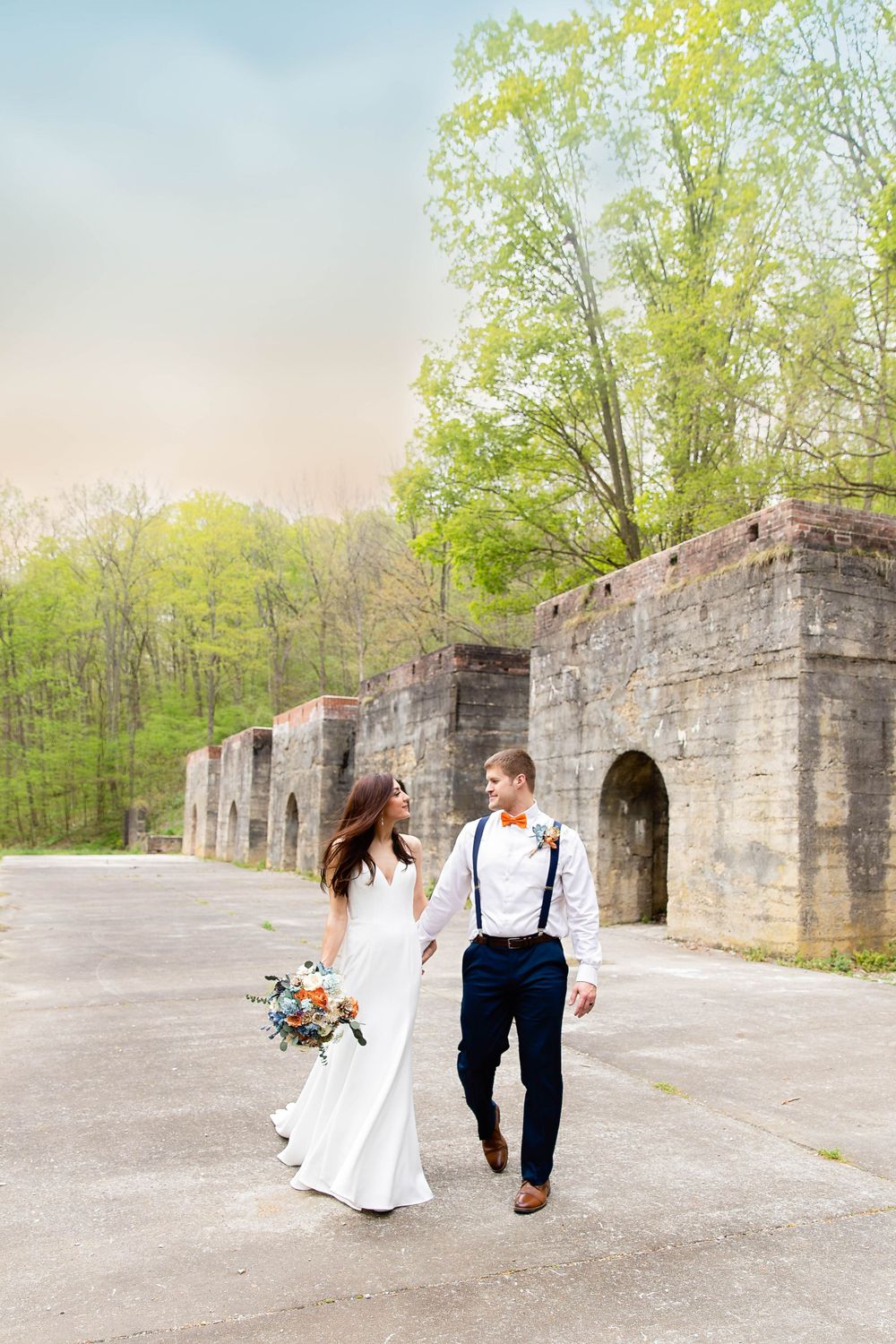A romantic wedding couple walks along stone ruins in a spring forest setting with blooming trees in the background.