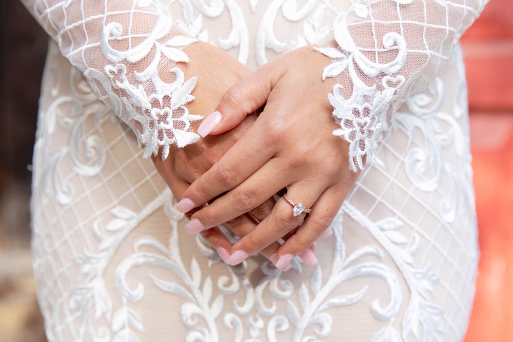 Elegant white lace wedding dress detail with engagement ring showing on clasped hands.