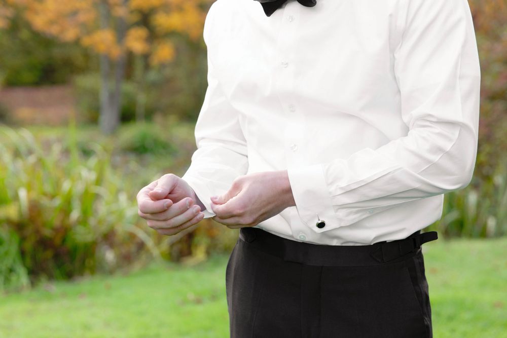 Close-up view of someone in formal attire adjusting their black bow tie against a blurred garden background.