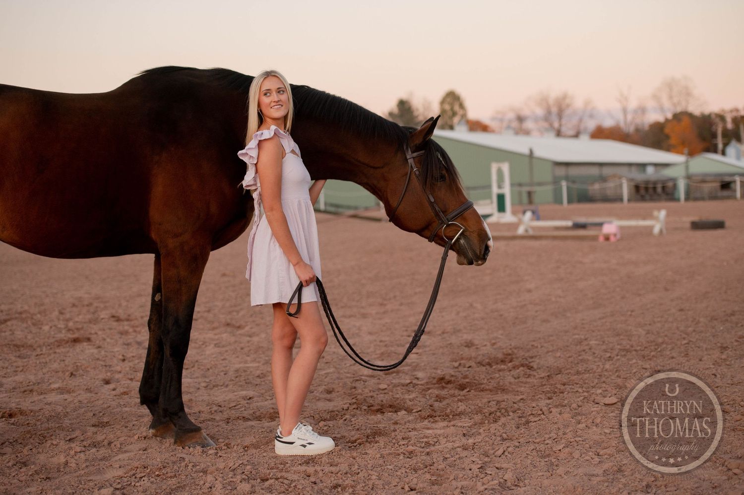 Someone in a white dress stands beside a dark horse in a sandy arena during sunset.