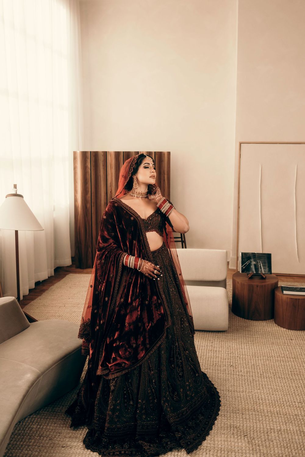 A person wearing a luxurious black and red velvet lehenga poses in an elegant interior setting.