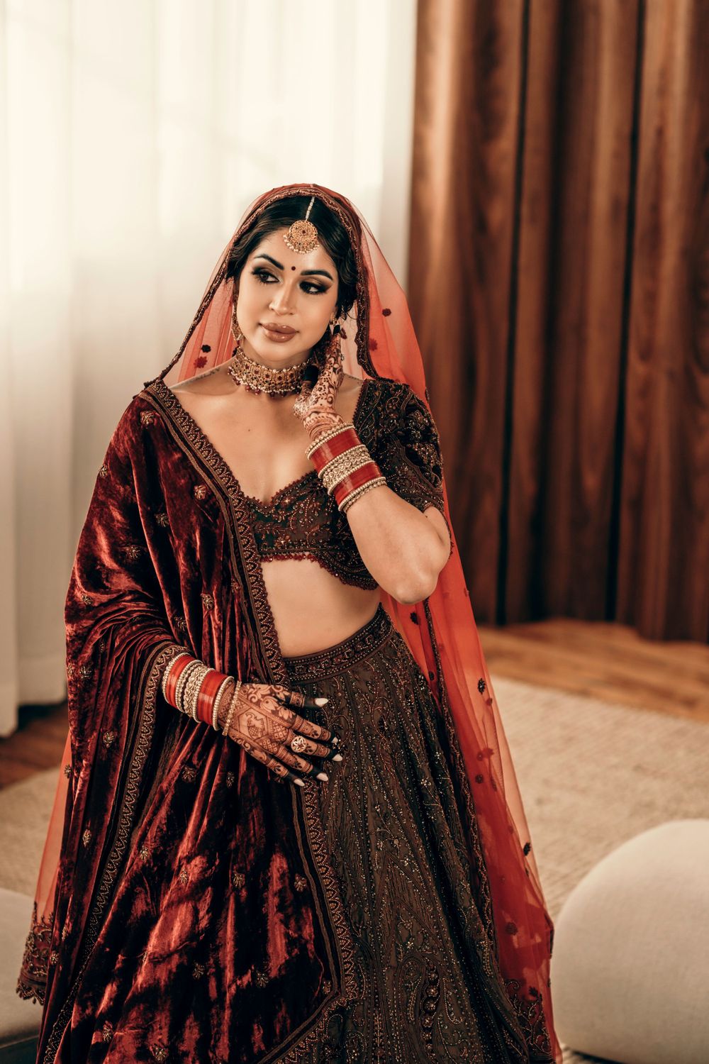 Bride in traditional red and burgundy Indian lehenga with ornate jewelry and henna poses indoors.