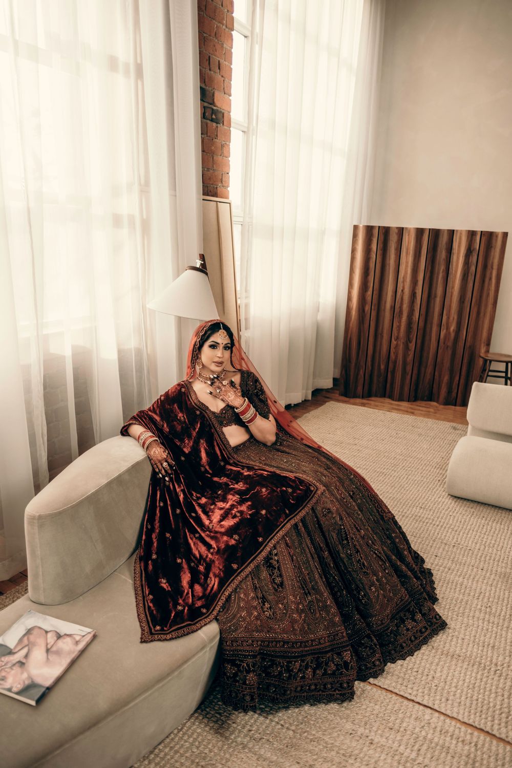 An elegant bridal portrait featuring a luxurious burgundy and black lehenga on a cream sofa with sheer curtains.