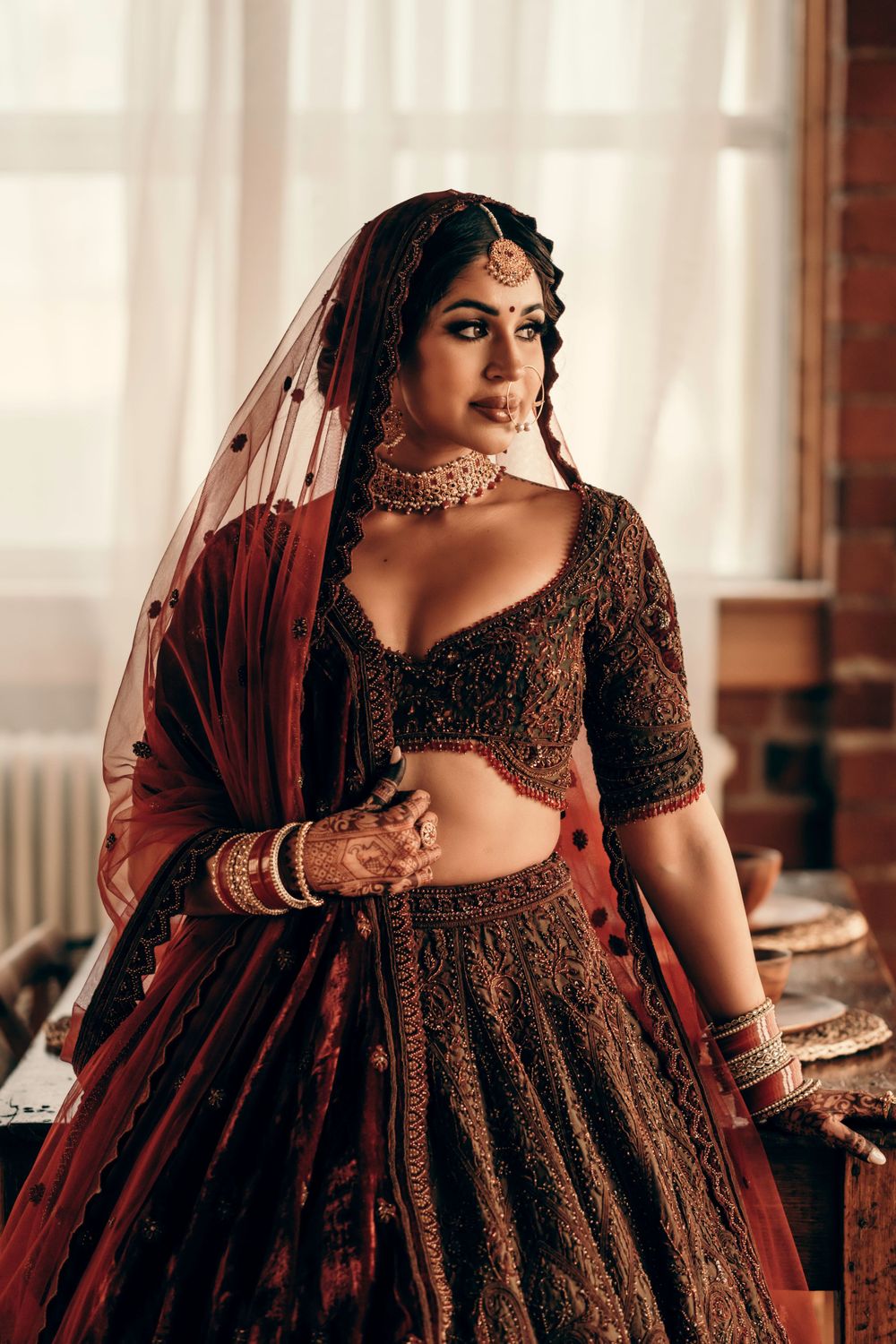 A traditional South Asian bridal outfit featuring an intricately embroidered brown lehenga and maroon veil with matching jewelry.