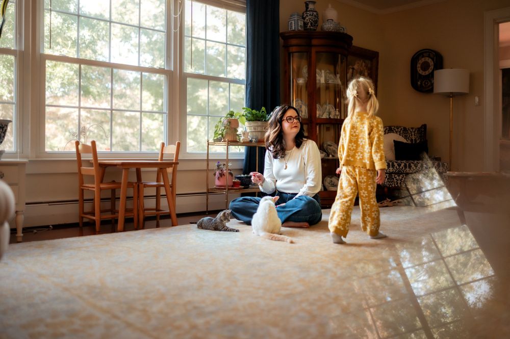 A golden doodle dog stands next to someone sitting cross-legged on a sunlit carpeted floor by large windows.
