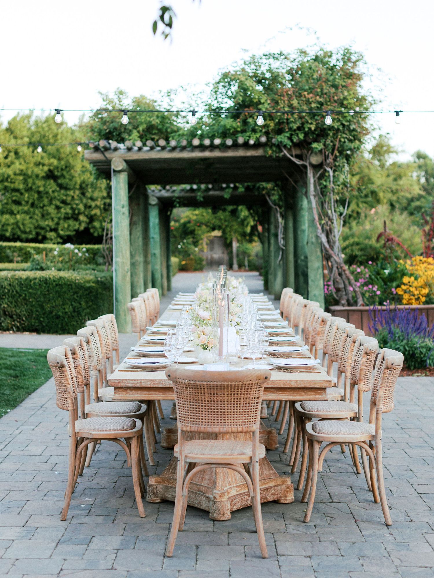 Elegant outdoor dining setup with rattan chairs and wood tables arranged in rows on a stone patio beneath garden pergolas.