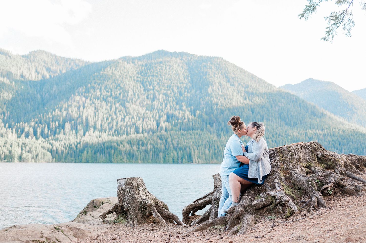 couple sitting on stump and kissing in front of scenic lake with mountains behind them