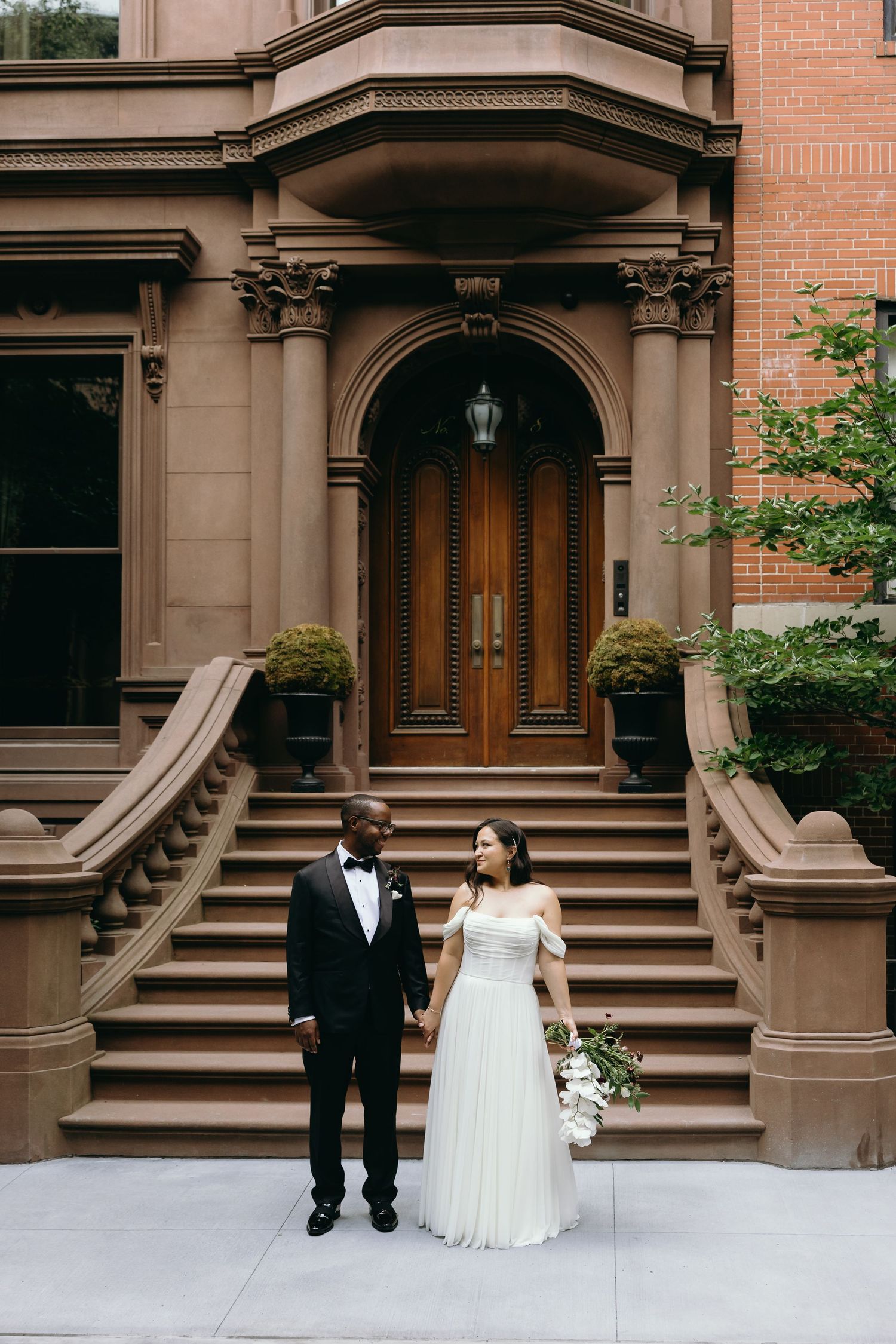 A couple in wedding attire stands on the steps of a historic brownstone building with ornate architectural details.