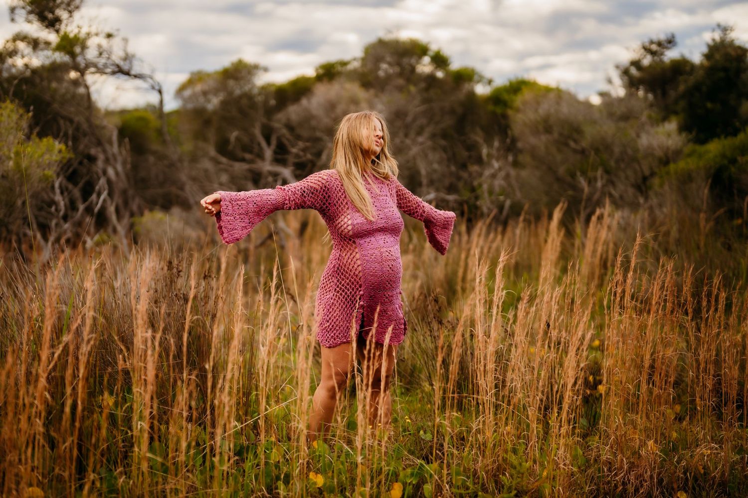 A pregnant woman in a pink dress stands with outstretched arms in a golden field of tall grass at sunset.