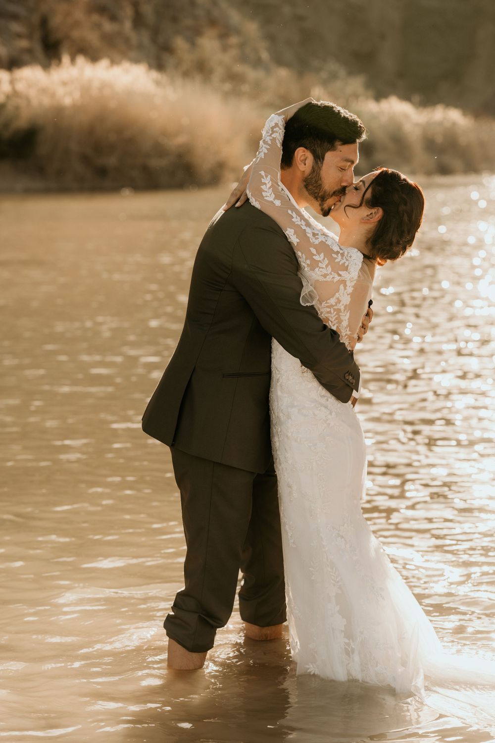 Couple in wedding attire standing knee-deep in river water during sunset.