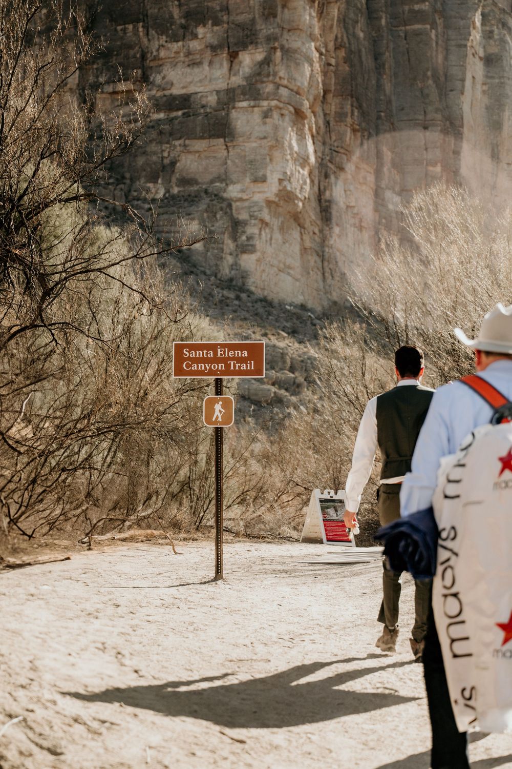 Desert hiking trail with warning signs and rocky cliffs in the background.