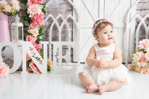 A baby in a floral dress sits on a white lattice bench surrounded by pink flowers in a garden setting.