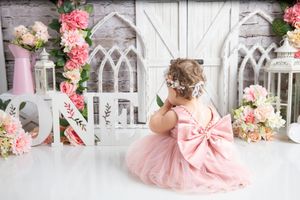 A pink tulle dress with bow detail is photographed against an ornate white backdrop with pink and peach floral arrangements.