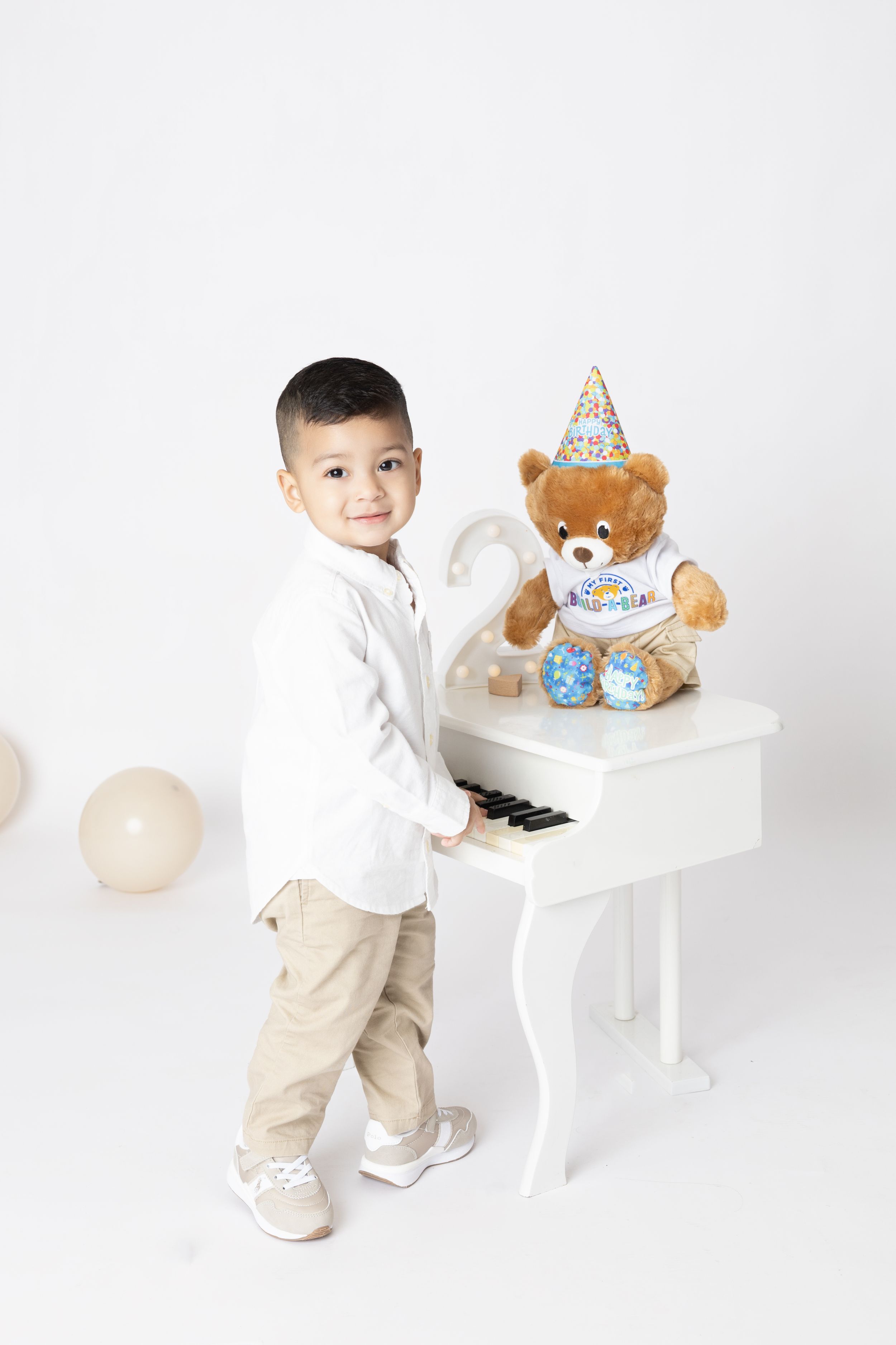 A child in white shirt and khaki pants stands next to a white piano with a teddy bear wearing a party hat on top.